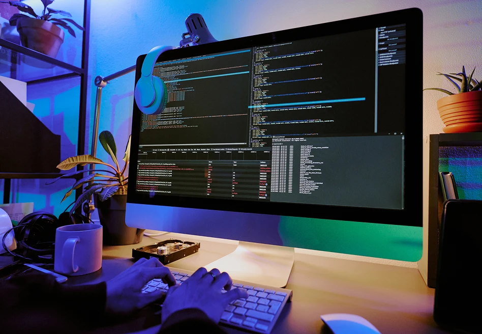Software developer working at desktop computer with code editor and system logs on large monitor, modern workspace at night
