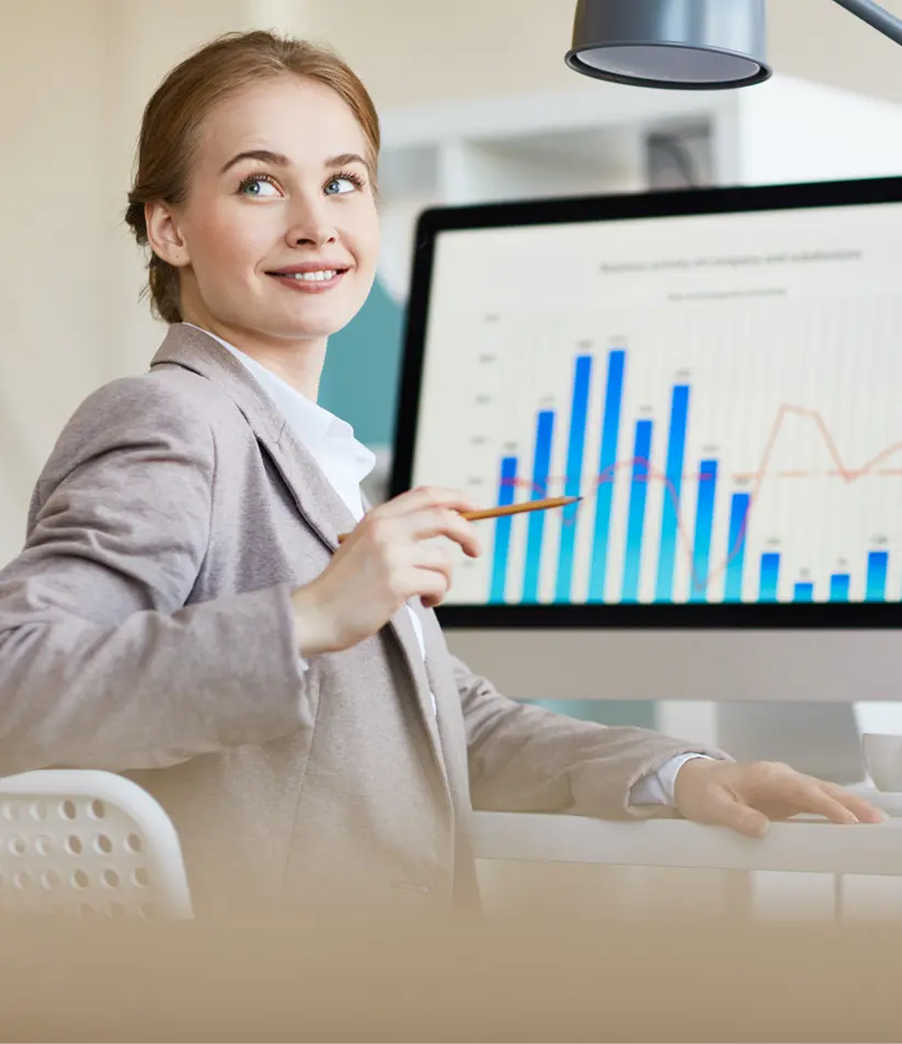 Smiling businesswoman holding a pen at a desk with a bar chart displayed on a monitor behind her