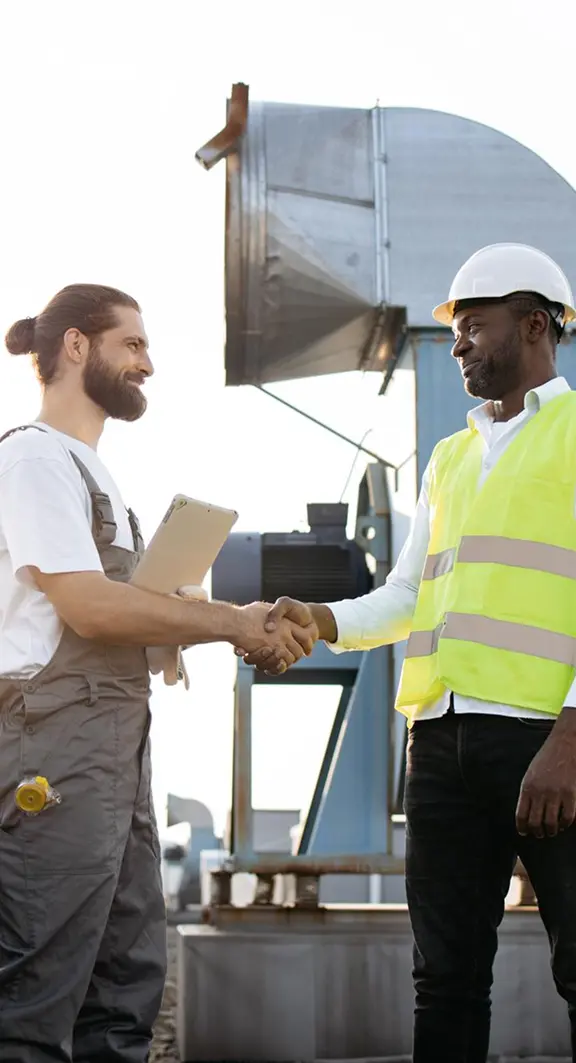 Two workers shaking hands on a rooftop near industrial HVAC equipment, one holding a tablet and one wearing a hard hat and high-visibility vest