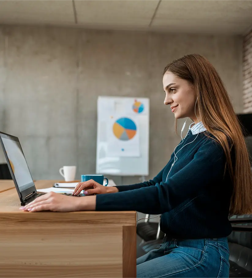 Young woman with earphones working on a laptop at an office desk with a pie chart on the wall behind her