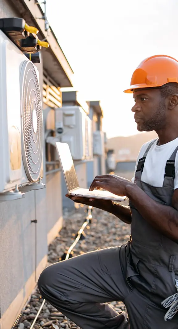 HVAC technician in an orange hard hat using a laptop while inspecting rooftop air conditioning units