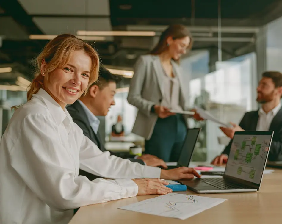 Smiling businesswoman at a conference table with colleagues working on laptops during an office meeting