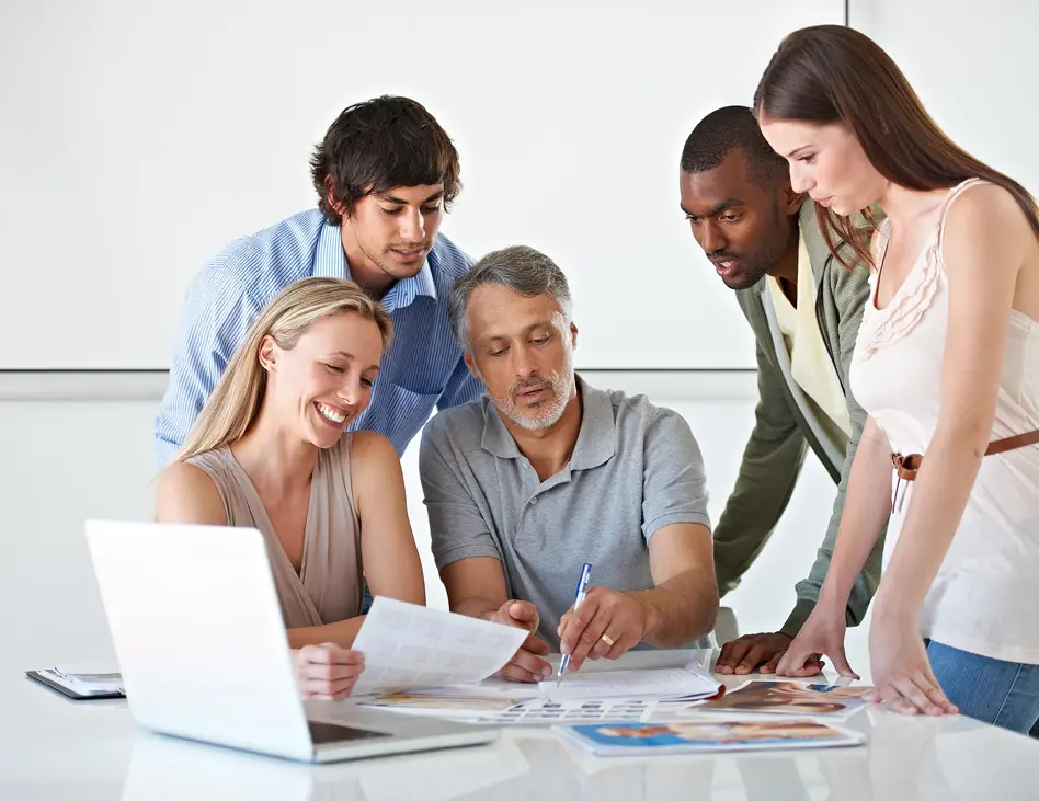 Diverse team of five colleagues gathered around a table reviewing documents and photos with a laptop